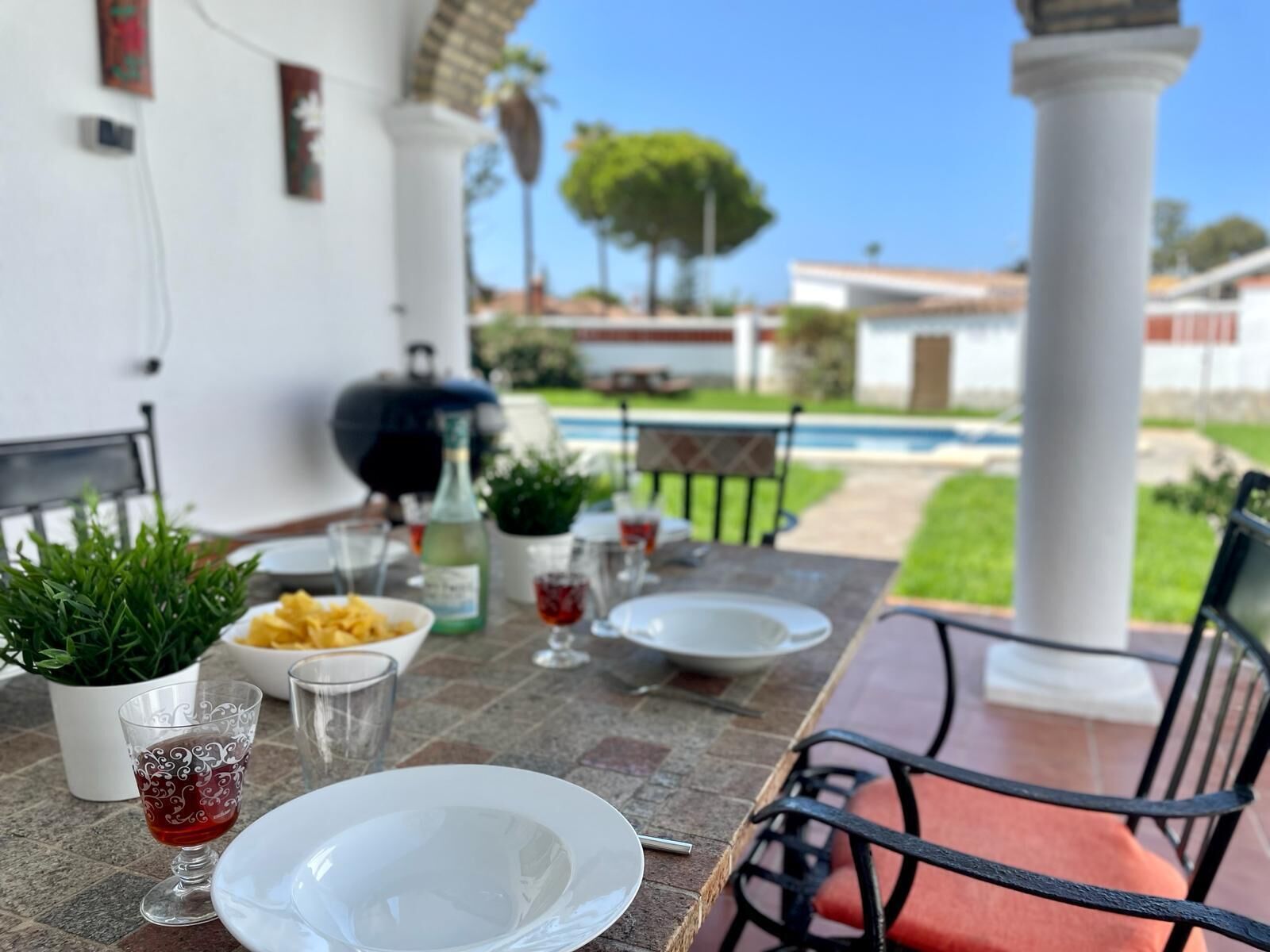 Un espace extérieur aménagé pour les repas, avec une table dressée pour deux personnes, un barbecue et une piscine en arrière-plan.