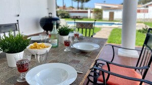 An outdoor dining area with a table set for two, a grill, and a pool in the background.