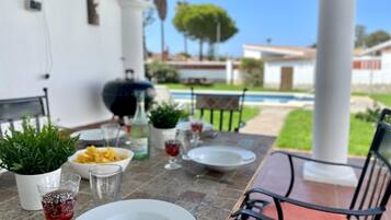 Un espace extérieur aménagé pour les repas, avec une table dressée pour deux personnes, un barbecue et une piscine en arrière-plan.