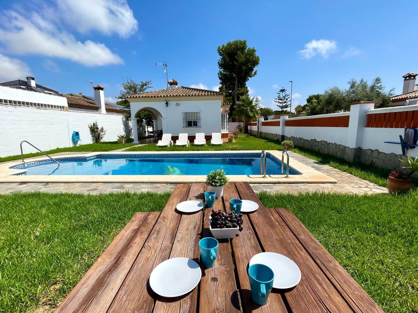 Un espace repas aménagé au bord de la piscine, avec une table en bois dressée pour deux personnes, une piscine et un bâtiment blanc en arrière-plan.