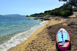 A beach with a surfboard on the sand, clear water, and a rocky shoreline. - Apartment in French Riviera with Pools (GRIMAUD)