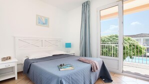 A bedroom with a bed, a nightstand, a lamp, a window with curtains, and a view of a balcony. - Apartment in French Riviera with Pools (GRIMAUD)