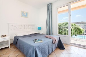 A bedroom with a bed, a nightstand, a lamp, a window with curtains, and a view of a balcony. - Apartment in French Riviera with Pools (GRIMAUD)