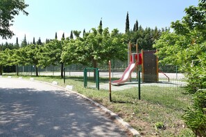 Ein Spielplatz mit roter Rutsche und Kletterwand, umgeben von Bäumen und einem Zaun.