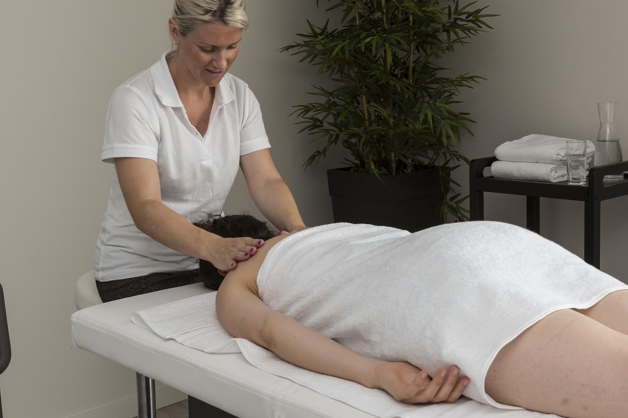 Eine Person erhält eine Massage in einem Raum mit Massagetisch, einer Zimmerpflanze und einem kleinen Tisch mit Handtüchern und einem Glas.