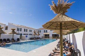 Una piscina con sillones de descanso y sombrillas de paja, rodeada de edificios blancos con contraventanas verdes.