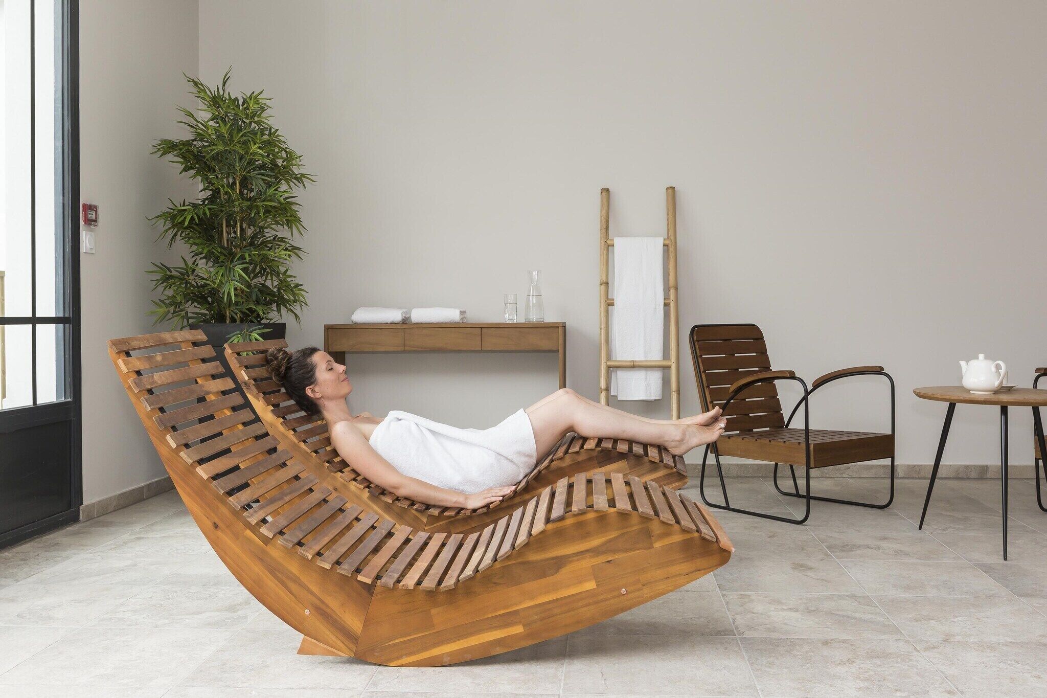 A person reclining on a wooden lounge chair in a room with a plant, a mirror, and a small table with a teapot.