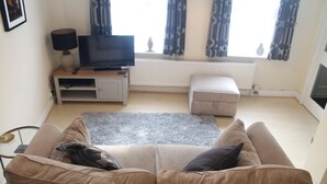 Een woonkamer met een beige bank, een flatscreen televisie op een houten dressoir, een grijs vloerkleed en een raam met patroon gordijnen.