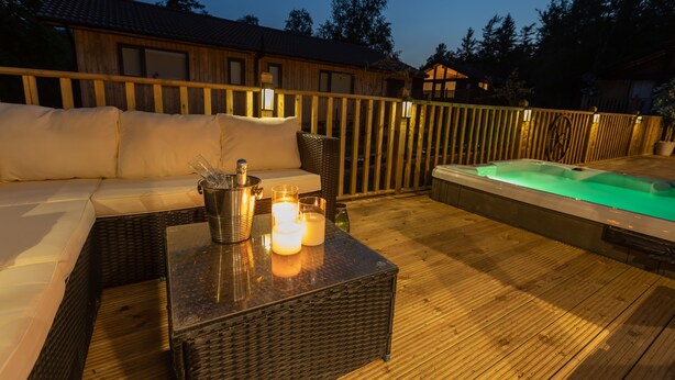 Un terrazzo in legno con un divano in vimini, una vasca idromassaggio e un tavolo con candele.
