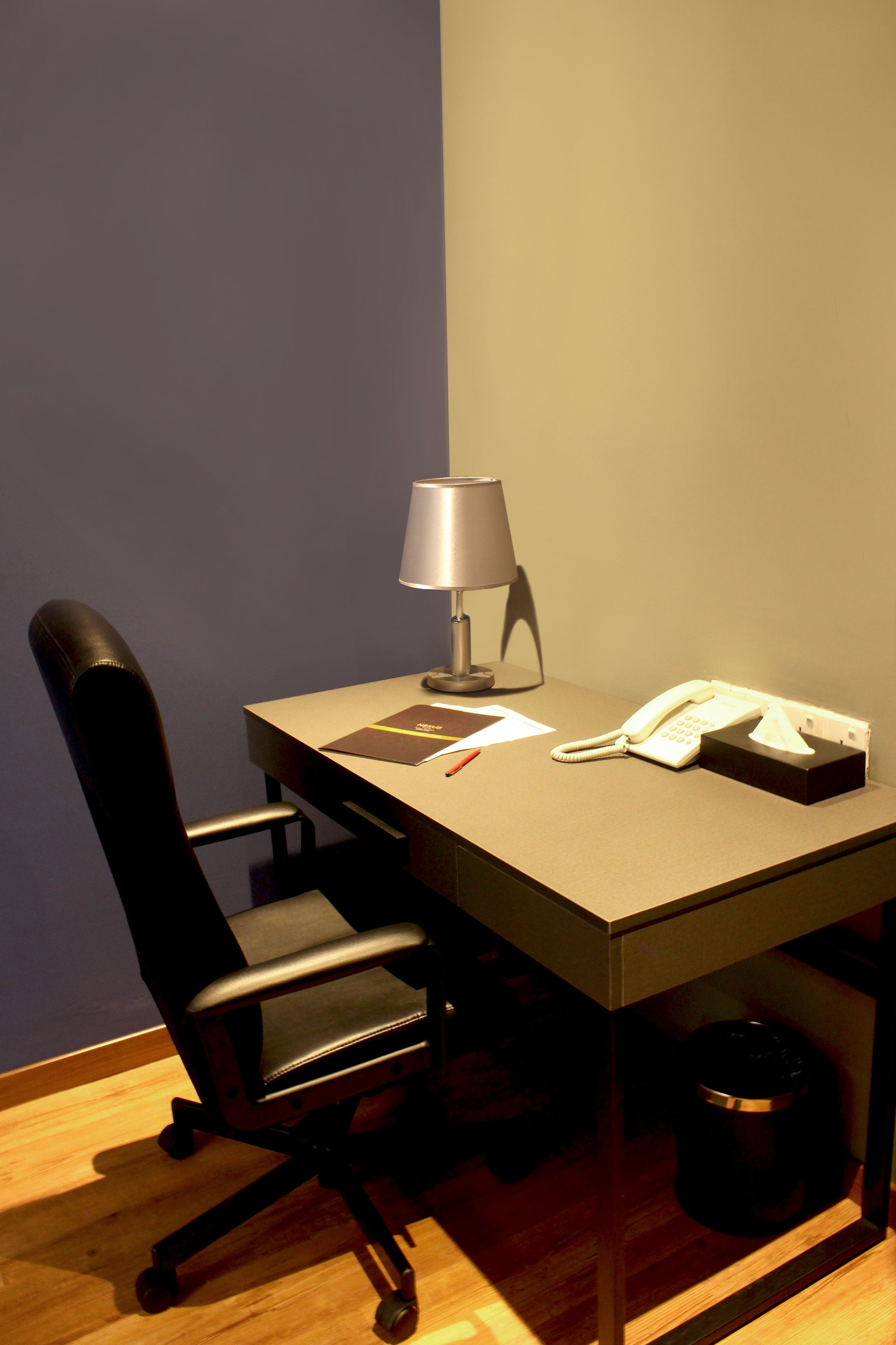 A small office space with a desk, chair, lamp, telephone, and a trash bin.