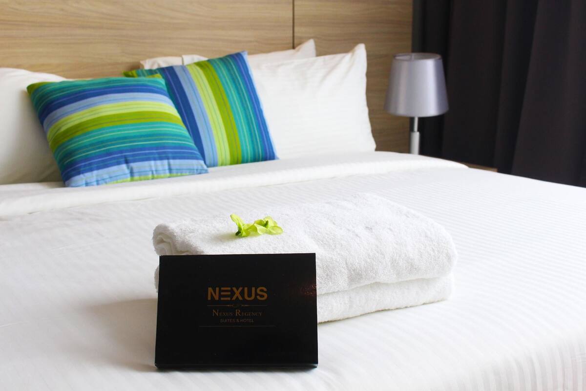 A neatly made bed with a white duvet, a folded white towel, and a black sign reading "NEXUS Nexus Regency Suites & Hotel".