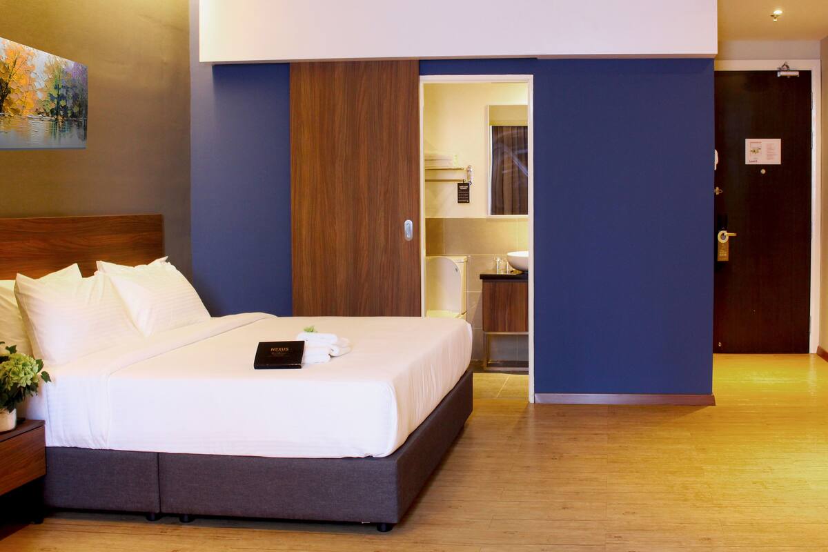 A hotel room with a large bed, a nightstand with a plant, a painting on the wall, and a bathroom visible through an open door.