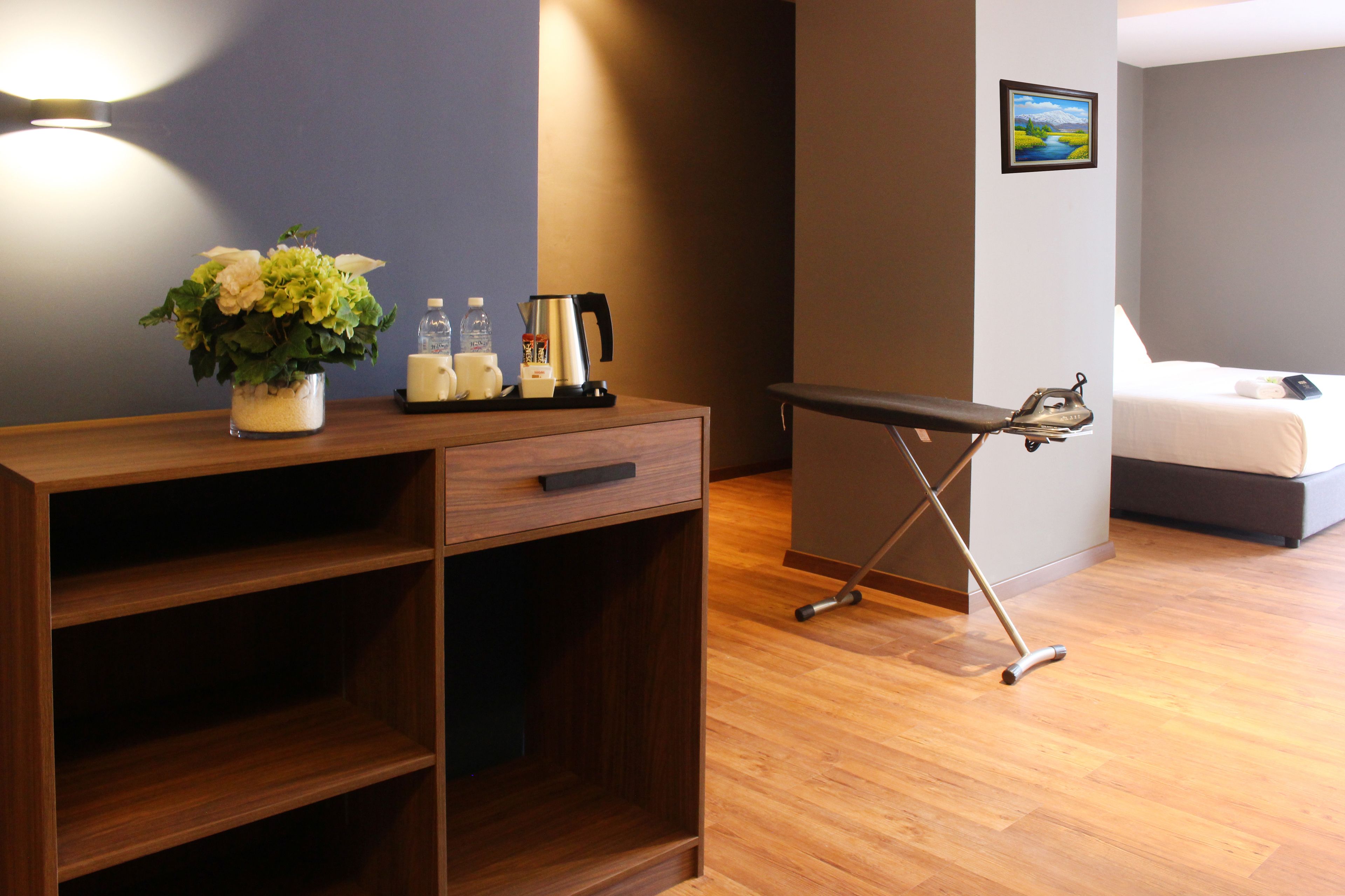A hotel room with a wooden desk, a small plant, a kettle, and a ironing board.