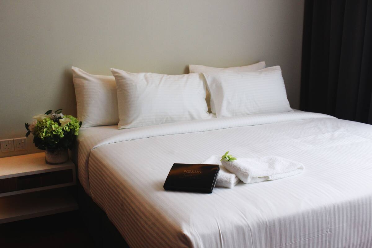 A neatly made bed with white linens, a black box labeled "NEXUS," and a small potted plant on a bedside table.