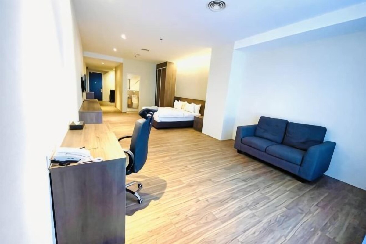 Ein Hotelzimmer mit Holzboden, Schreibtisch mit Stuhl, blauem Sofa und Bett mit Kissen.