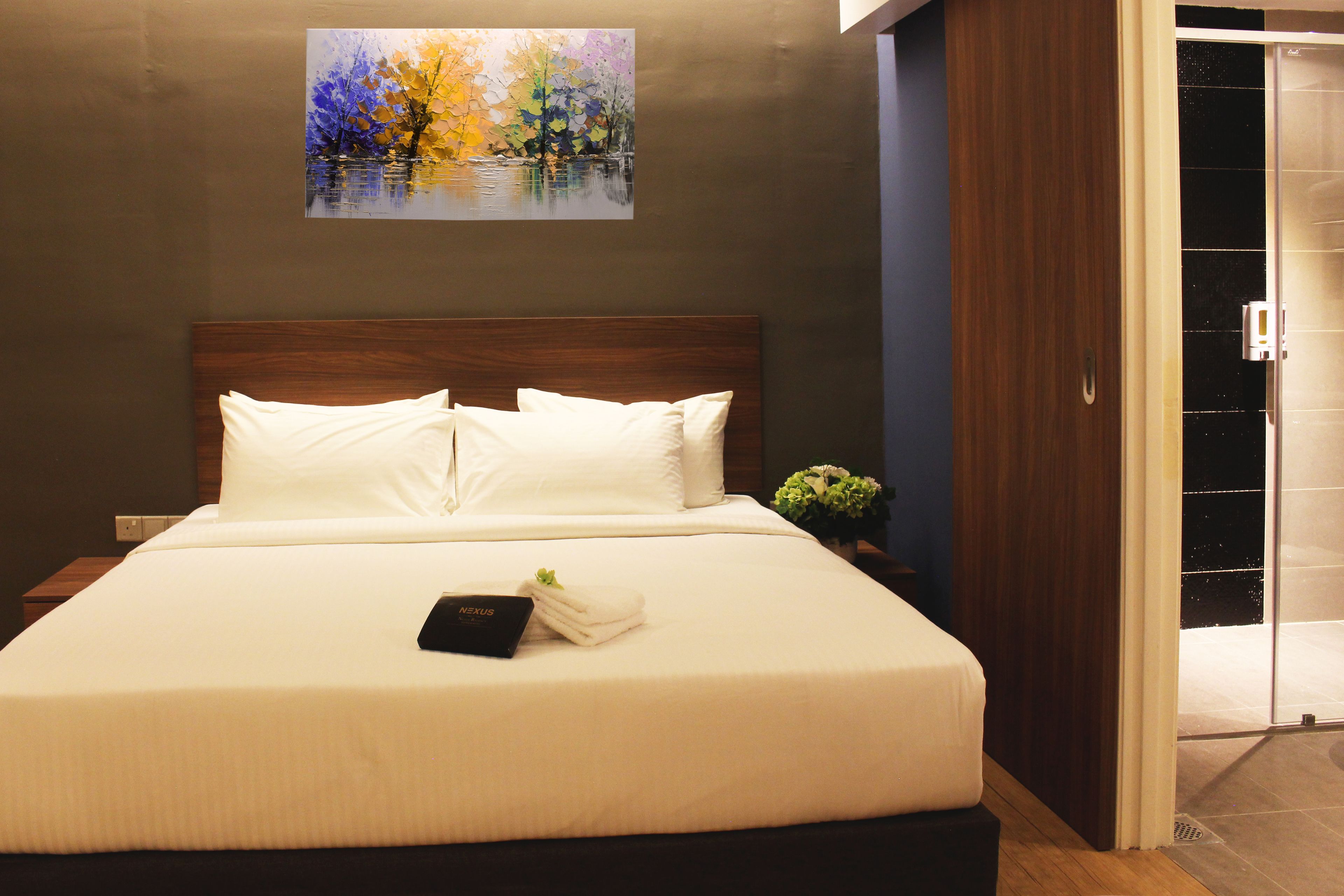 A neatly made bed with white linens and pillows, a small bedside table with a book and a folded towel, and a painting of trees on the wall.