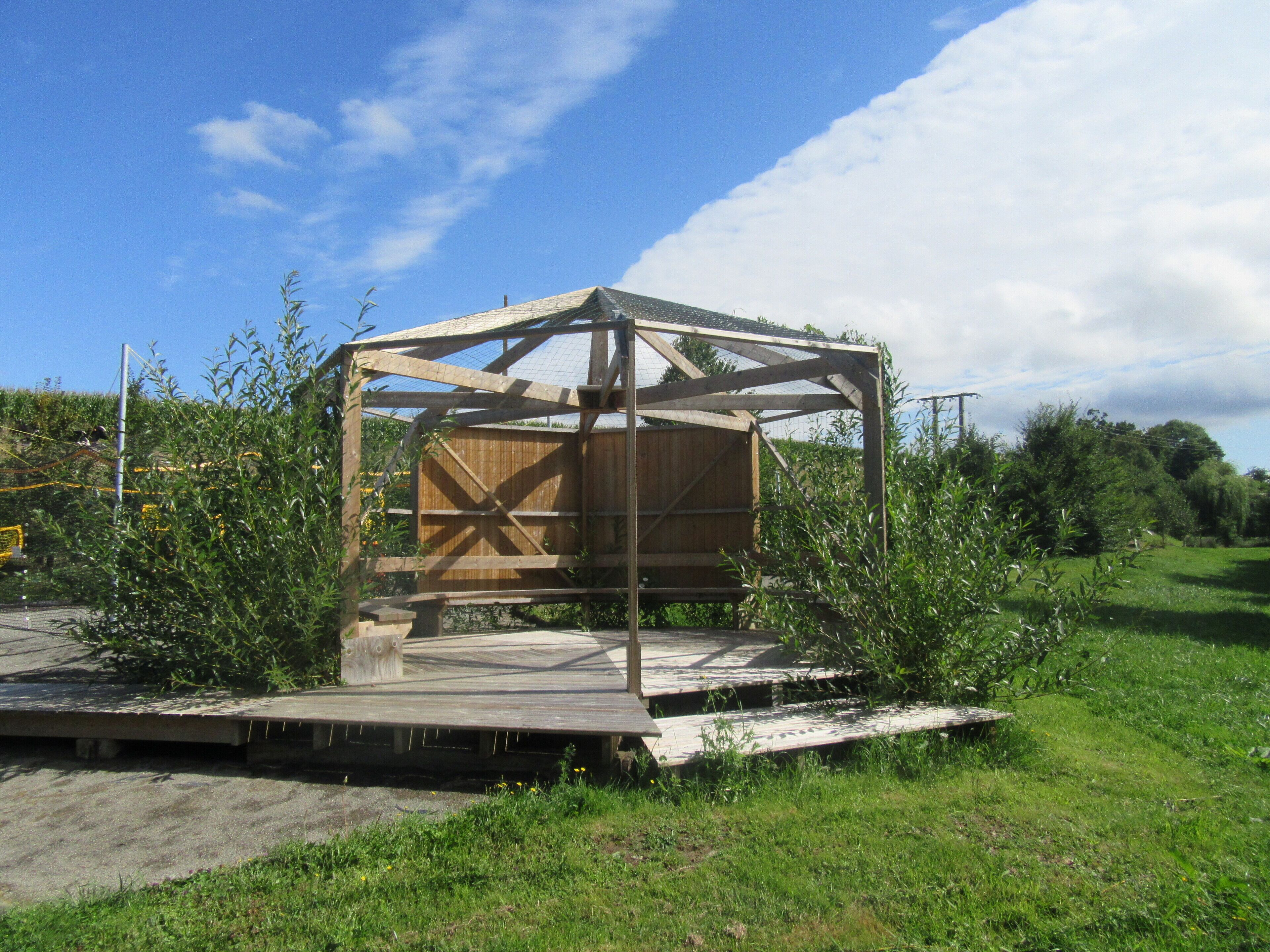 A wooden structure with a metal frame, surrounded by greenery and a clear sky.