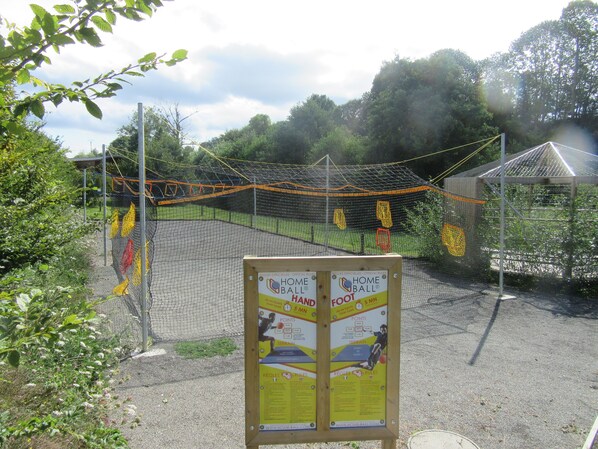 Ein Sportplatz mit einem durch ein Netz abgetrennten Torbereich, umgeben von Bäumen und einer Holzkonstruktion.
