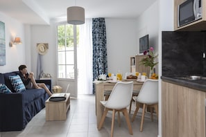 Ein modernes Wohnzimmer mit einer Couch, einer Essecke mit Stühlen, einer Küche inklusive Mikrowelle und einem großen Fenster.
