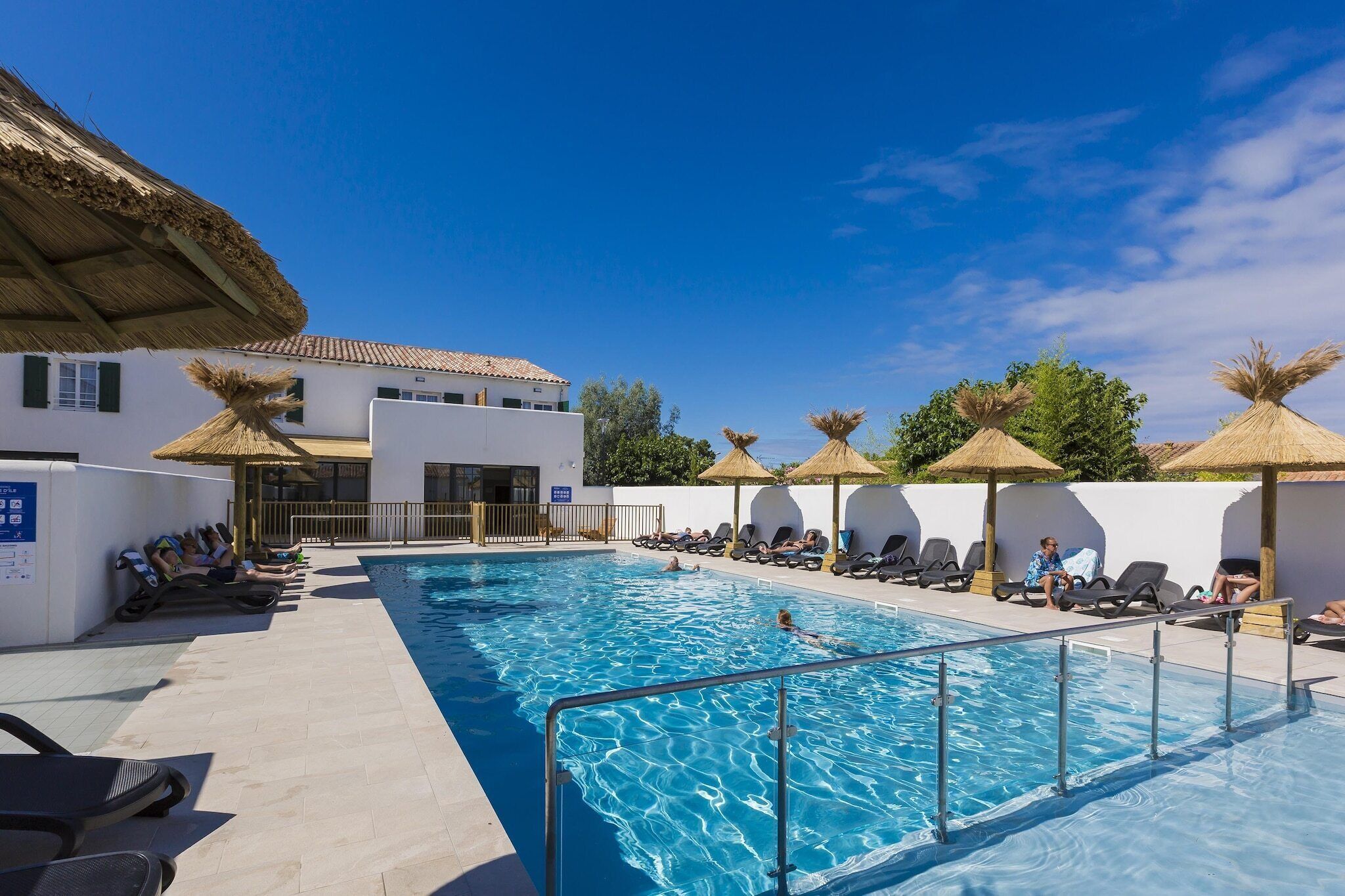 Une piscine avec des chaises longues et des parasols, un bâtiment doté d’un toit en tuiles et un ciel bleu azur.