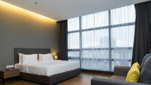 Una habitación de hotel moderna con una cama grande, una mesita de noche, un sofá y un amplio ventanal con cortinas.