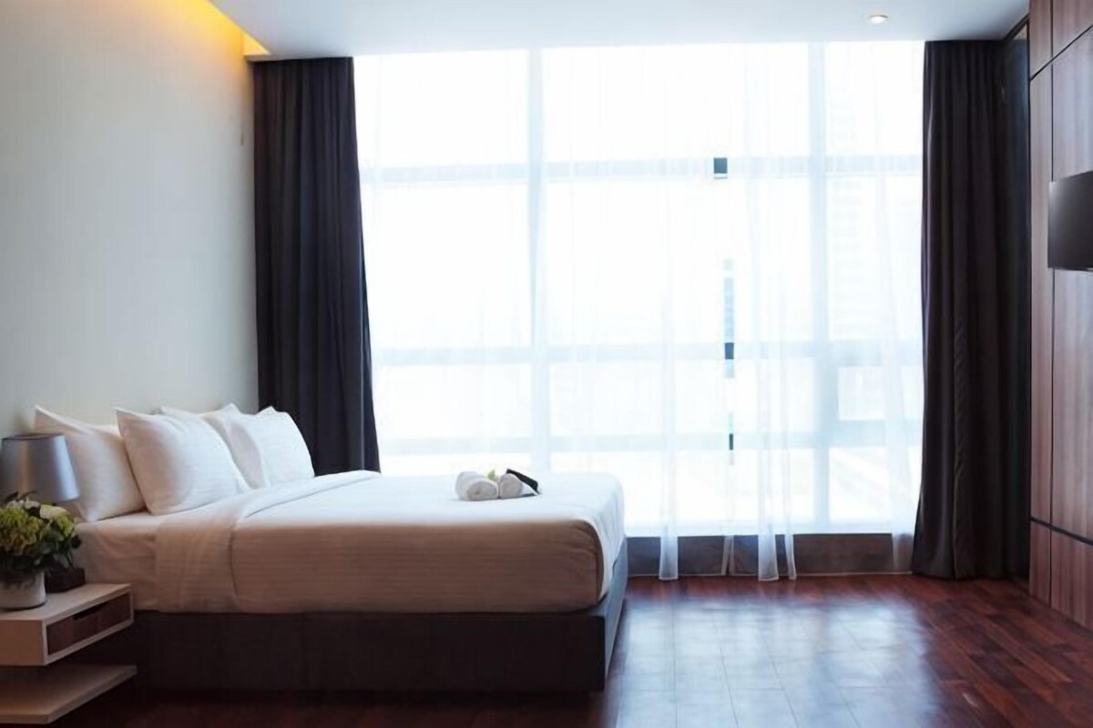A modern hotel room with a large bed, a bedside table with a lamp, and a view through sheer curtains.
