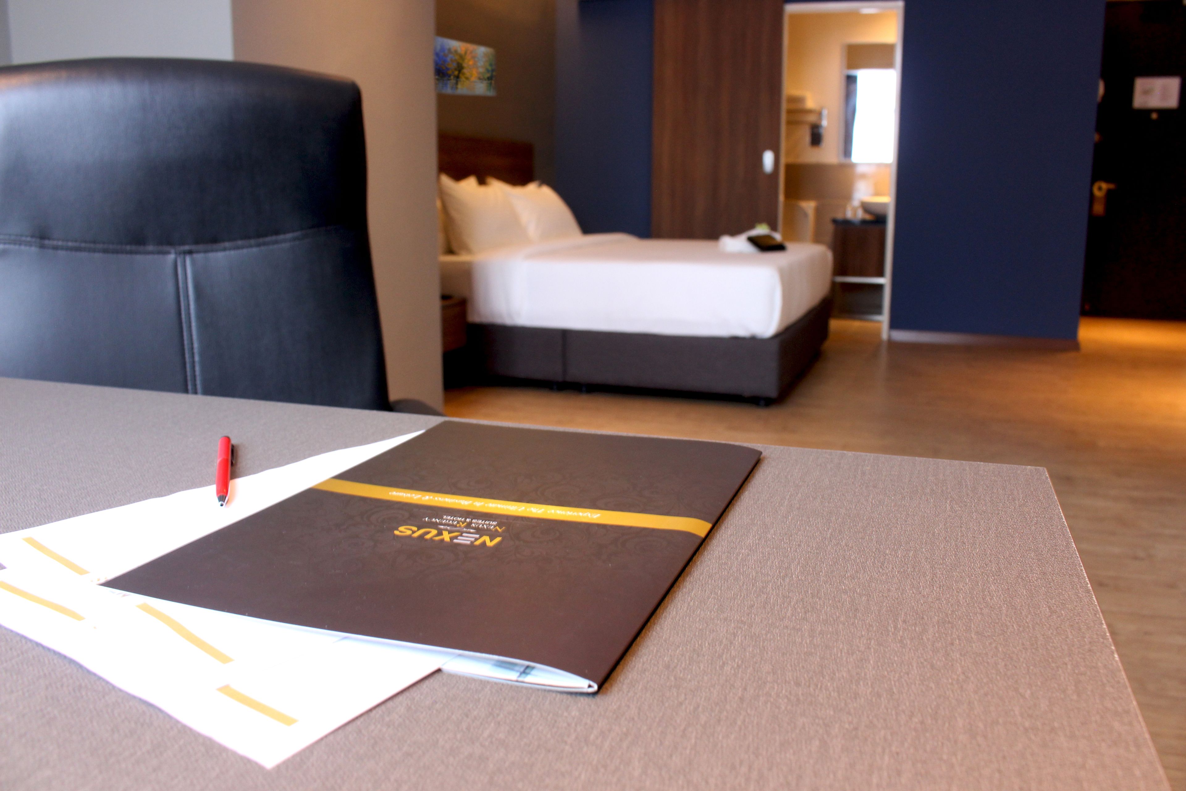 A hotel room with a bed, a desk with a notebook and pen, and a door.