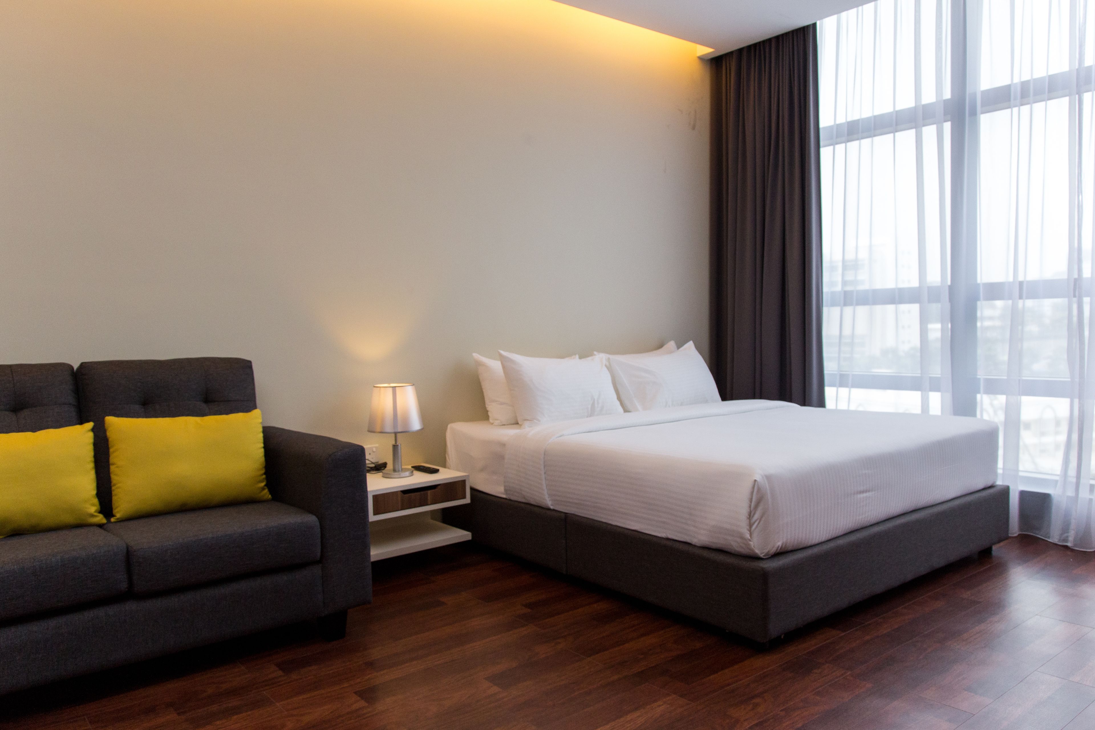 Una habitación de hotel moderna con una cama grande, un sofá con cojines amarillos, una mesita de noche con lámpara y vistas amplias a través de grandes ventanales.