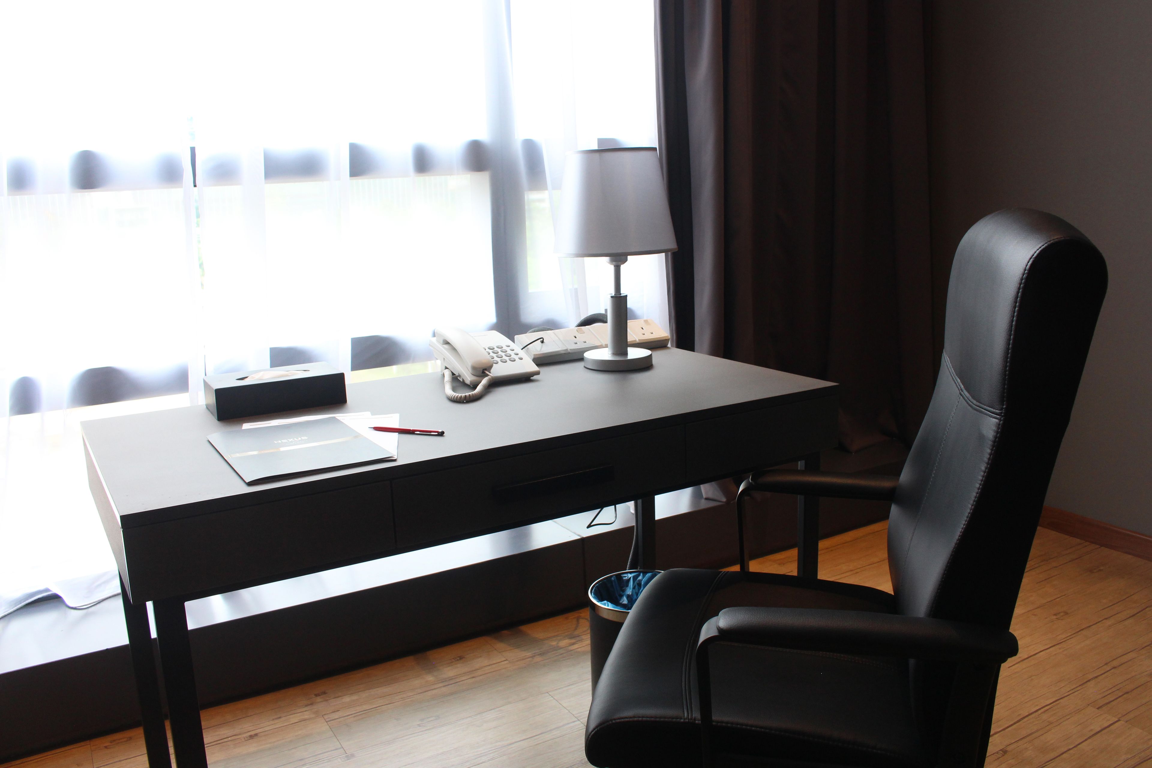 A hotel room with a desk, a chair, a telephone, a lamp, and a window with curtains.