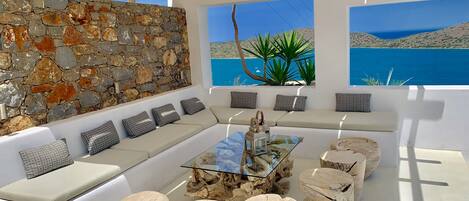 A sunlit outdoor seating area with a stone wall, a glass-top coffee table, and a view of the sea.