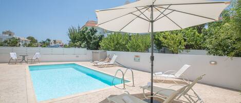 A swimming pool with lounge chairs and a large umbrella, surrounded by a tiled patio and greenery.