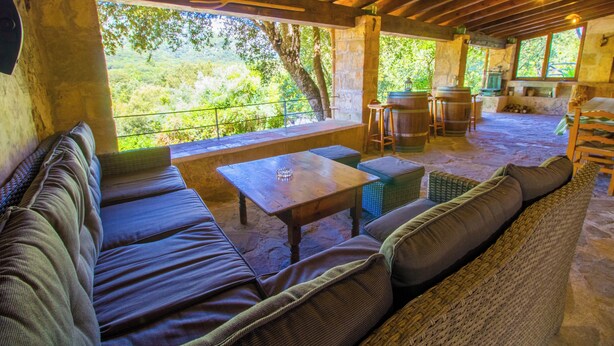 Patio esterno con tetto in legno, pareti in pietra e vista su un paesaggio verdeggiante. Si notano un tavolo in legno, una panca imbottita e una sedia in vimini.