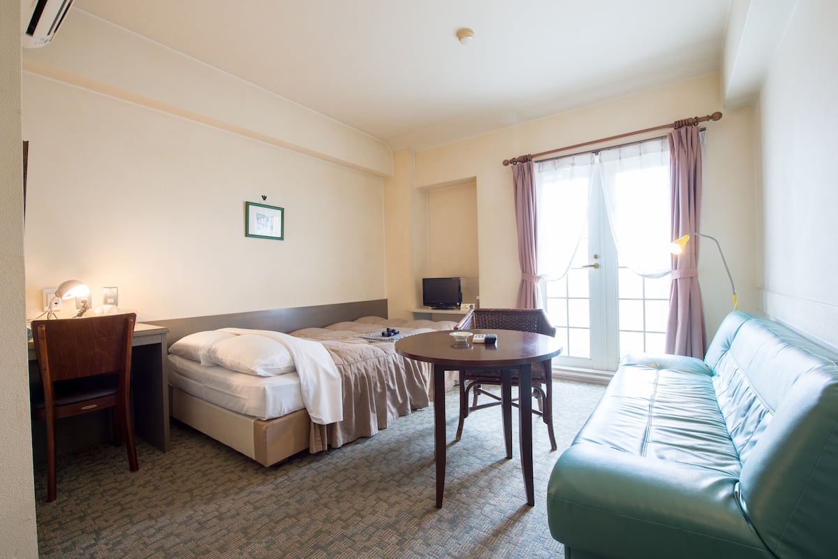 A hotel room with a bed, a desk with a chair, a small table, a couch, and a TV.