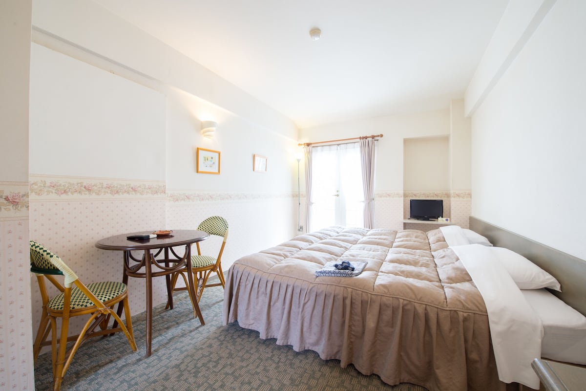 A hotel room with a bed, two chairs, a small table, a television, and a window with curtains.