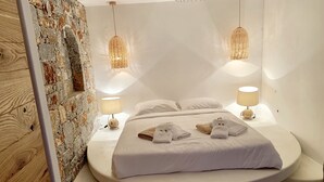 A modern bedroom with a stone wall, a curved bed, two bedside lamps, and neatly folded towels. - EXCEPTIONAL VILLA FACING THE SEA: VILLA INFINITY  (ELOUNDA)