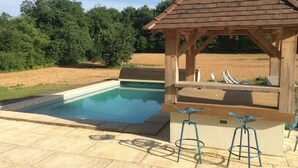 Un kiosque en bois doté d’un toit incliné, une piscine et deux tabourets de bar au design original.
