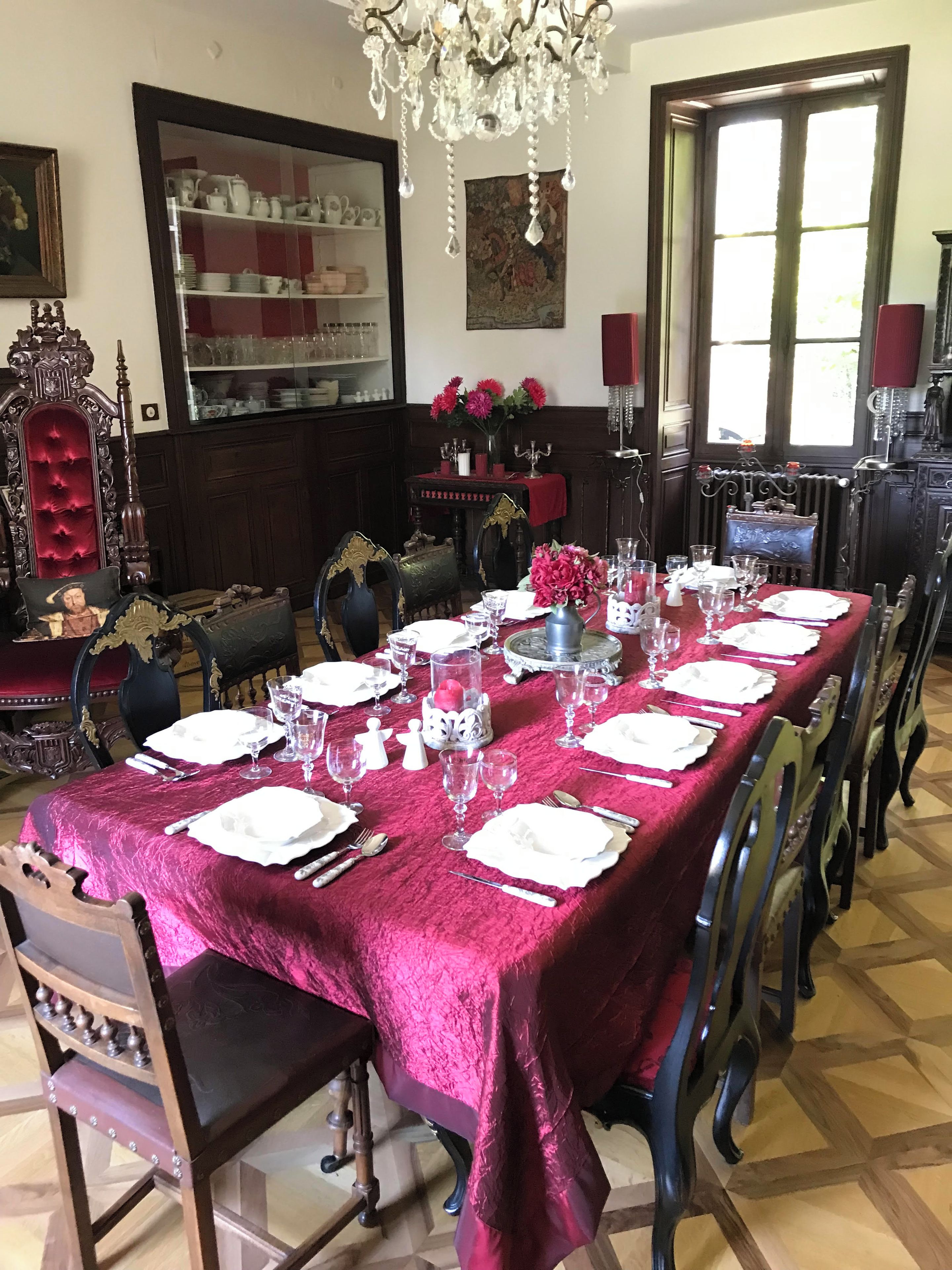 Une salle à manger avec une longue table dressée pour un repas, entourée de chaises richement décorées et surmontée d’un lustre.