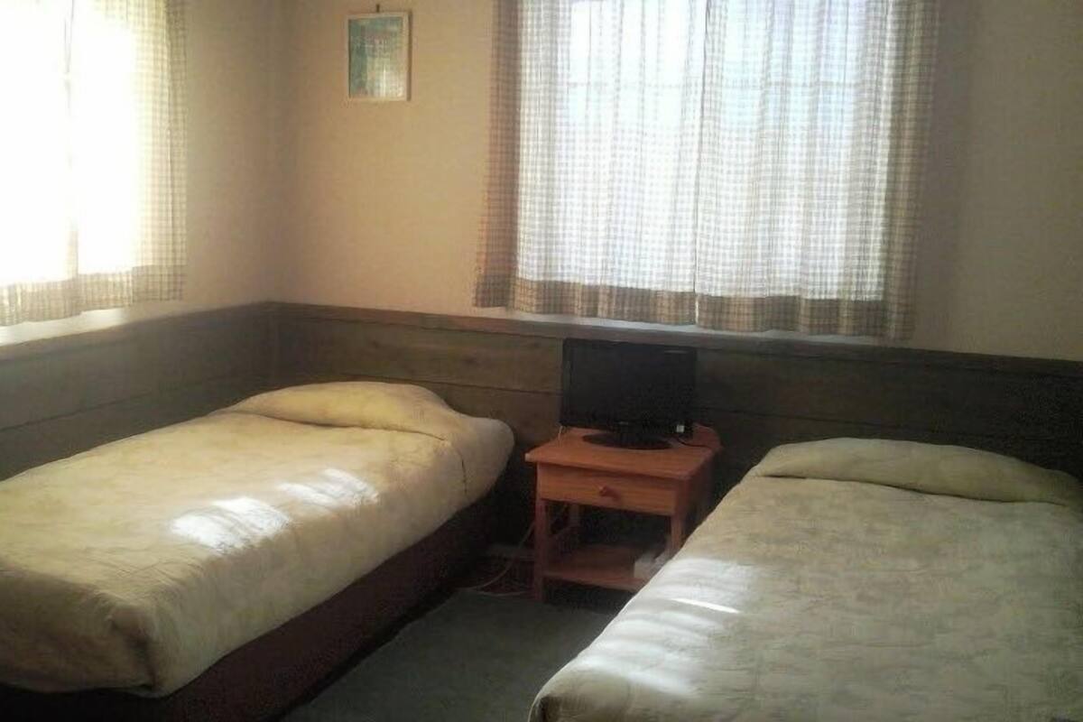 A room with two single beds, a wooden nightstand with a television, and a window with sheer curtains.