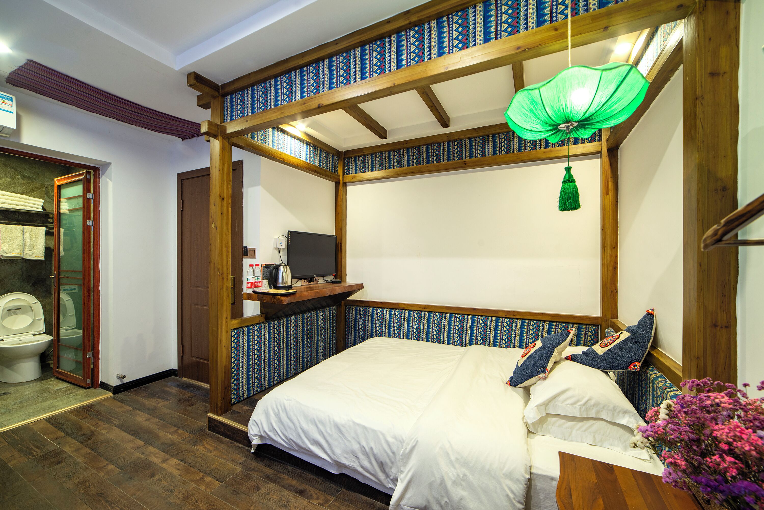 A room with a wooden ceiling, a bed with white bedding, a small table with a television, and a green hanging light fixture.