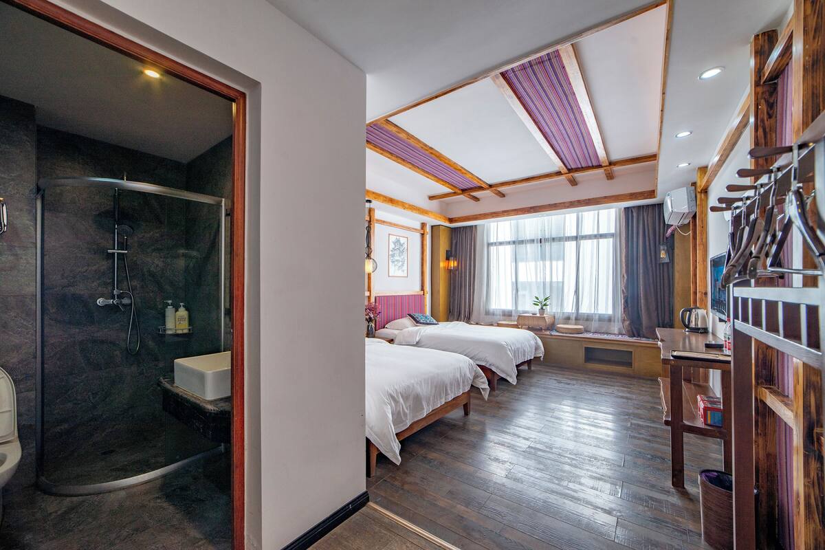 Ein Hotelzimmer mit einer Glaskabinedusche, Holzboden, zwei Betten, einem Holzregal mit Utensilien und einem Vorhangfenster.