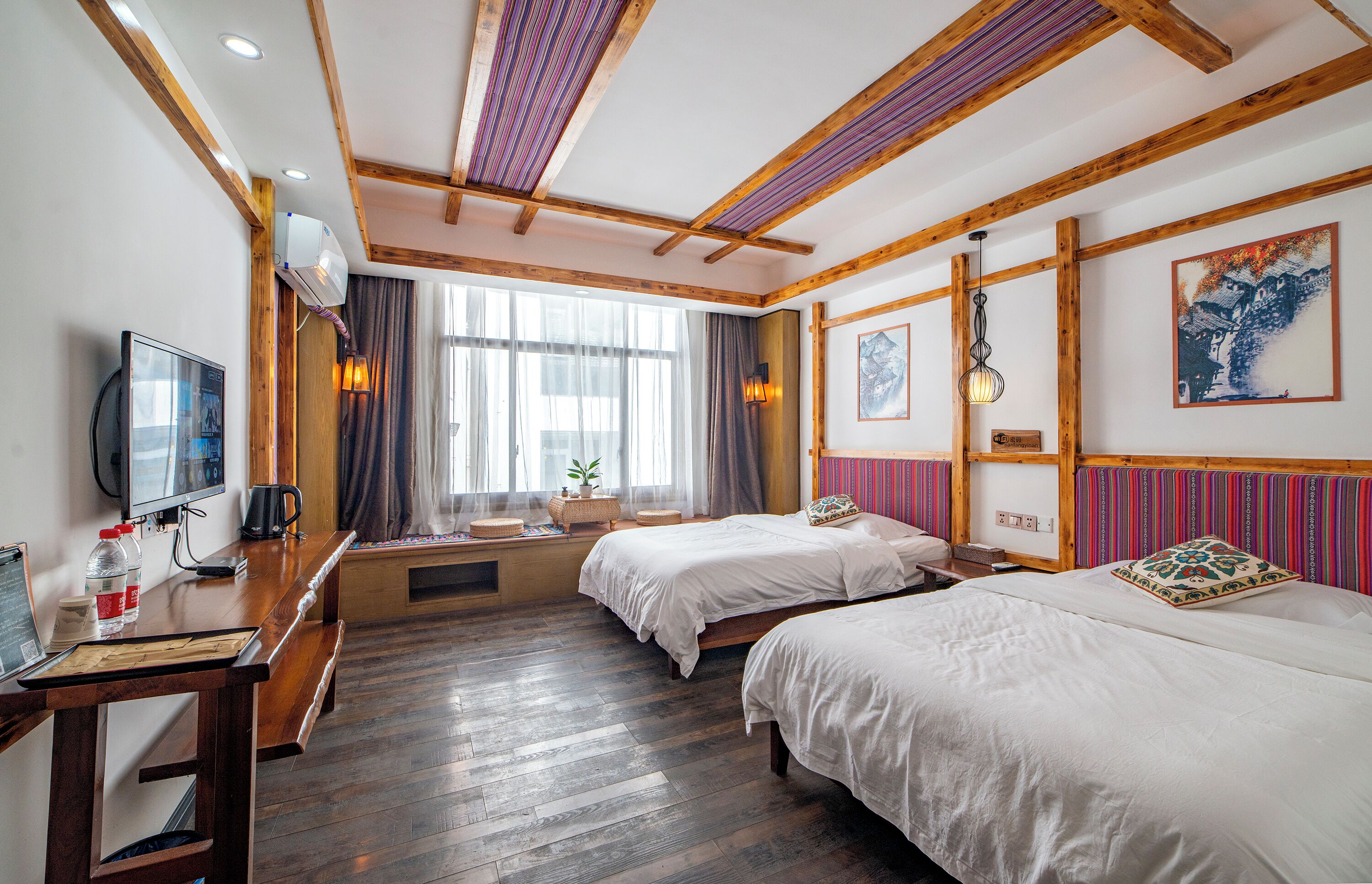 A hotel room with two beds, wooden beams on the ceiling, a TV, a desk with a computer, and a window with curtains.