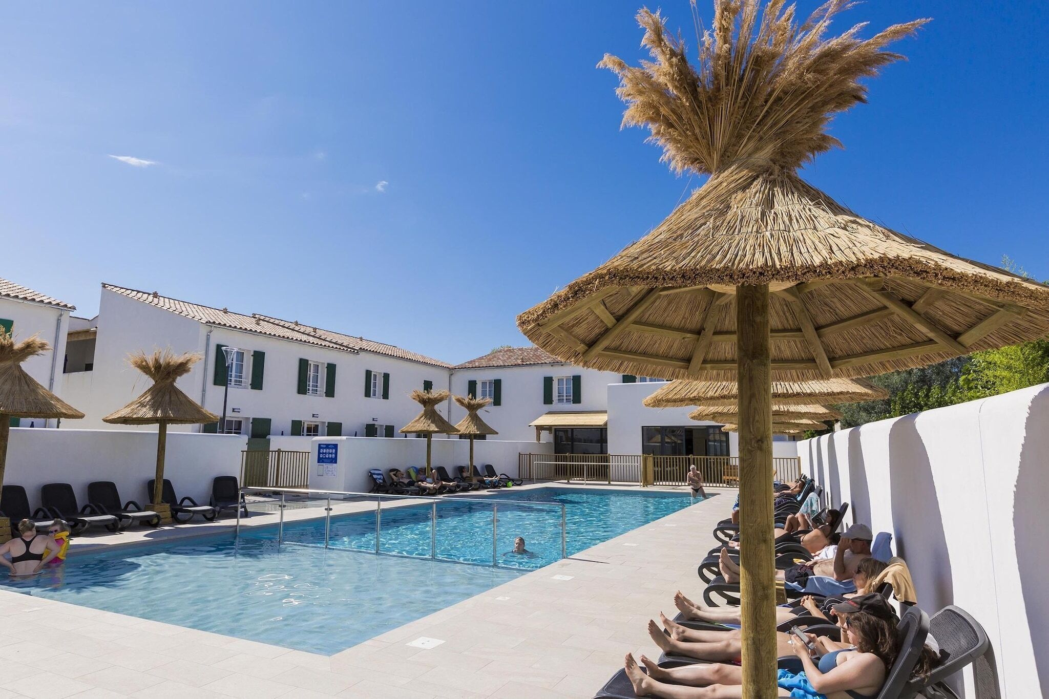 Una piscina amb gandules i sombrerets de palla, envoltada d'edificis blancs amb persianes verdes.