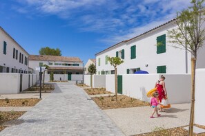 A pathway leading to a row of white buildings with green shutters, a clear sky, and a few trees.