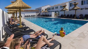 Ein Poolbereich mit Liegestühlen und Sonnenschirmen, ein klares, blaues Schwimmbecken sowie im Hintergrund ein Gebäude mit weißen Wänden und grünen Fensterläden.
