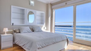 Een slaapkamer met een groot bed, een nachtkastje, een spiegel en uitzicht op de zee door een groot raam.
