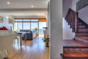 A spacious living area with a wooden floor, a sofa, a dining table, and a view of the sea through large windows.