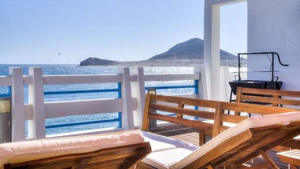 Een houten terras met lounge fauteuils en een barbecue, met uitzicht op een heldere blauwe zee en een bergachtig landschap.