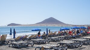 Een strand met rijen ligbedden en blauwe parasols, een berg op de achtergrond en mensen in het water.