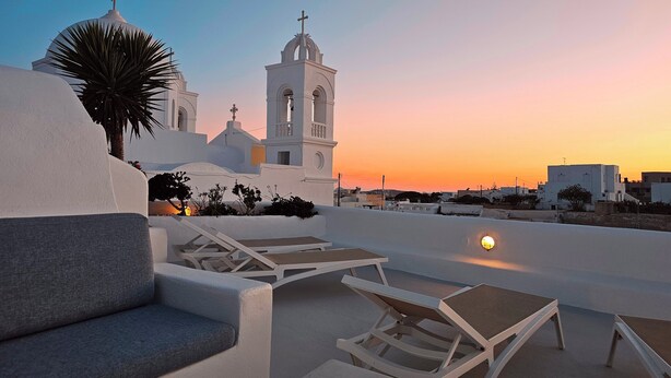 A rooftop terrace with white lounge chairs and a view of a white church with a bell tower against a sunset sky. - Traditional Village House 'Alba Lily' with heated plunge pool (Megalochori)