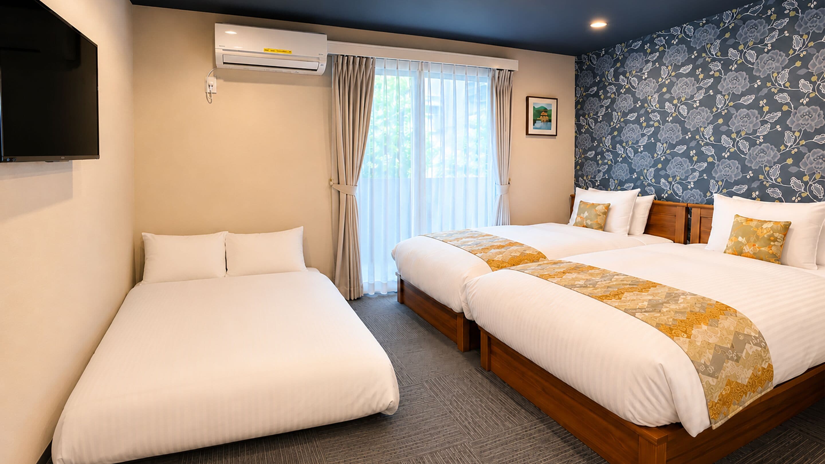 A hotel room with two beds, a flat-screen TV, an air conditioning unit, a window with curtains, and a floral wallpaper.